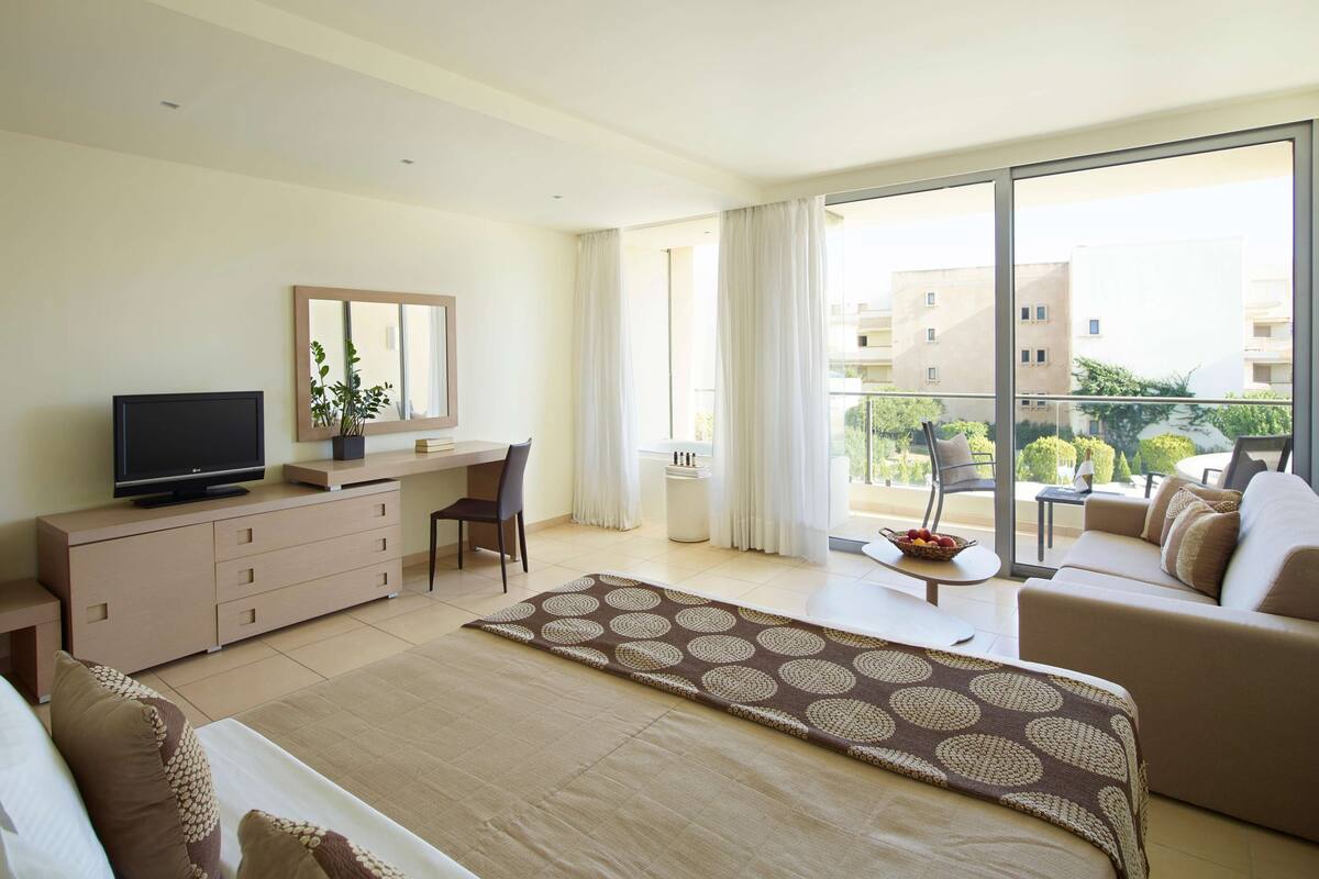 Habitación de hotel moderna con una cama grande, un escritorio con televisor, una silla, un sofá y un balcón con vistas a edificios y árboles.