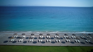 Vista aérea de una playa con filas de tumbonas y sombrillas, un mar azul transparente y montañas a lo lejos.