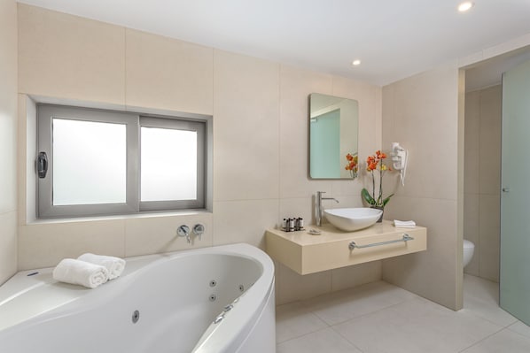 A modern bathroom with a large bathtub, a vanity with a sink, and a window. - The Ixian Grand & All Suites - Adults Only (Rhodes)