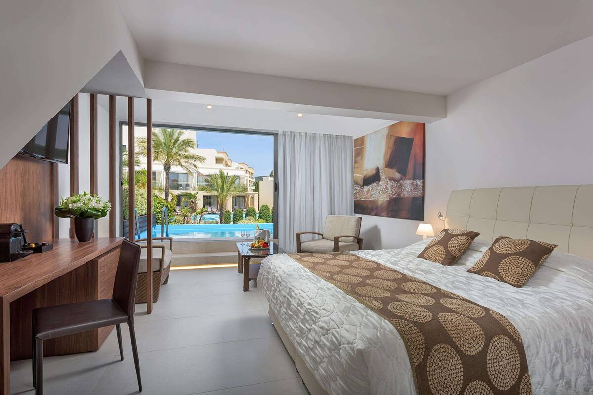 Habitación de hotel moderna con cama, escritorio, silla, televisión y vistas a una piscina y edificios.