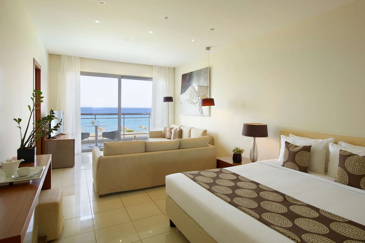 Habitación de hotel con una cama grande, un sofá, un escritorio con una silla, una mesita y vistas al mar a través de amplios ventanales.