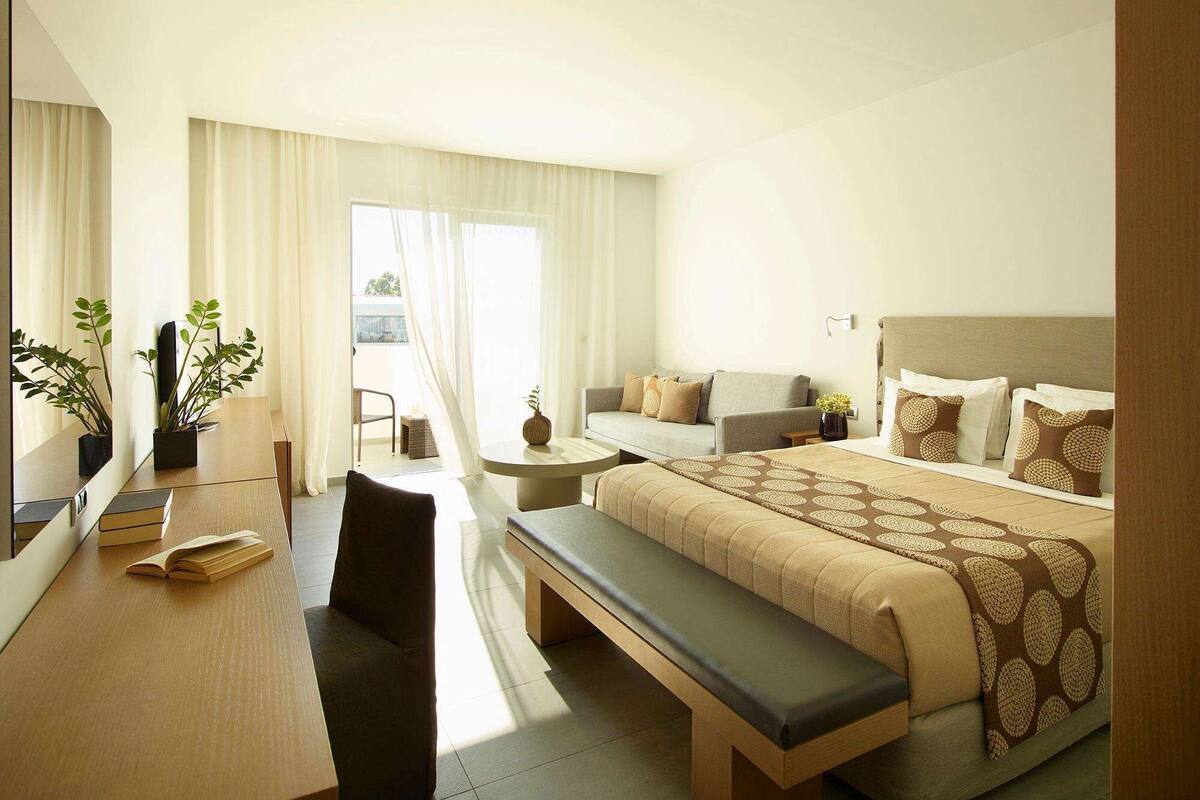 Una habitación de hotel con cama, escritorio, silla, banca, sofá y una mesita.