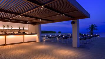 An outdoor dining area with tables and chairs, a bar area with a counter and plants, and a view of the ocean at dusk.