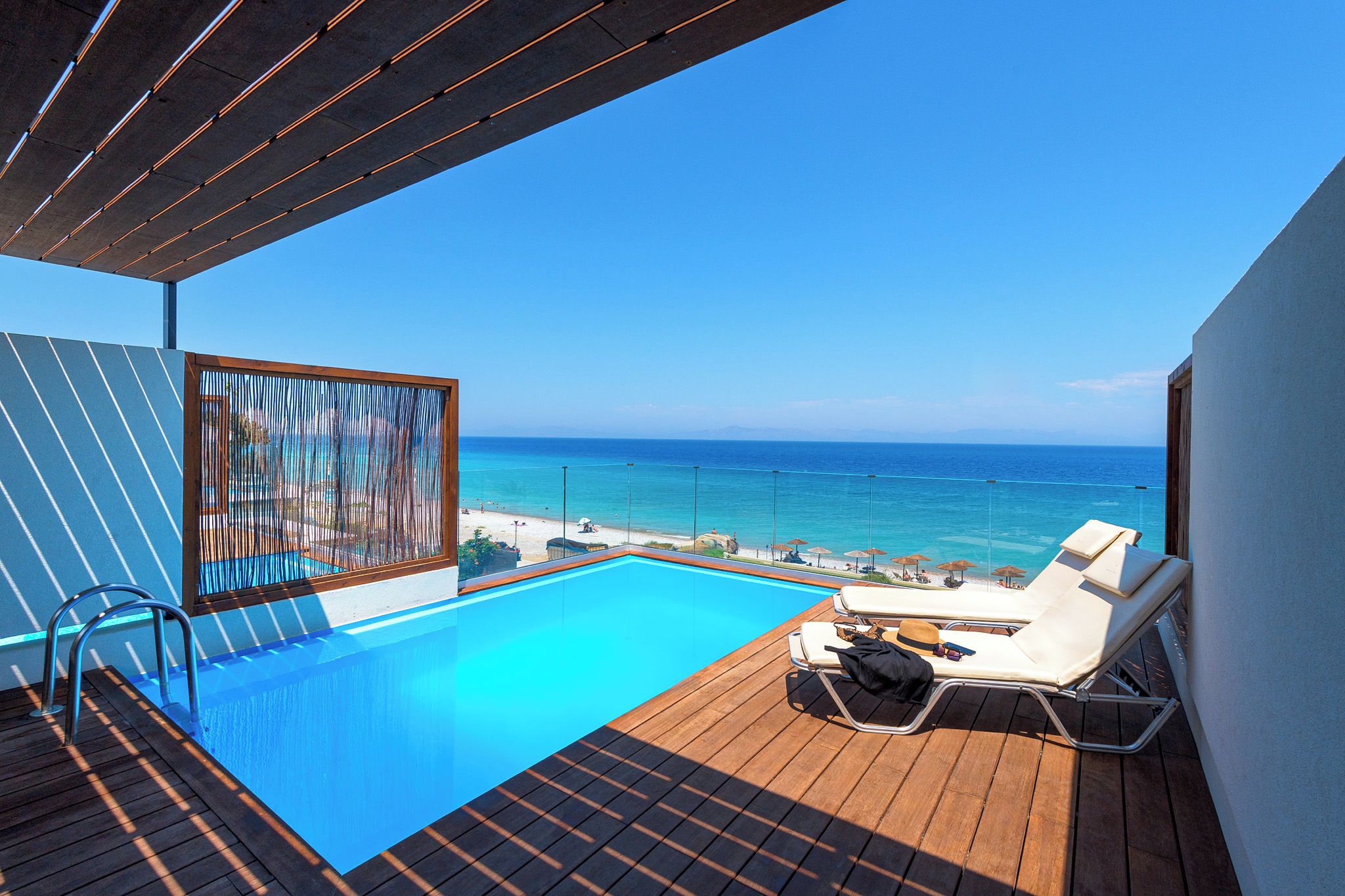 Zona junto a la piscina con vistas al mar, una terraza de madera y sillones de descanso.