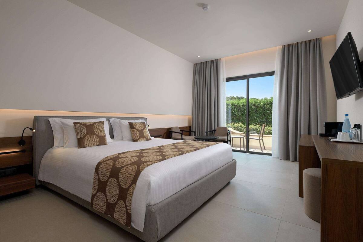 Habitación de hotel moderna con una cama grande, un escritorio, un televisor de pantalla plana y un balcón con vistas a la vegetación.