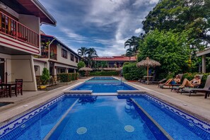 Una piscina con borde de baldosas, rodeada de sillones de descanso y sombrillas, con edificios y árboles al fondo.