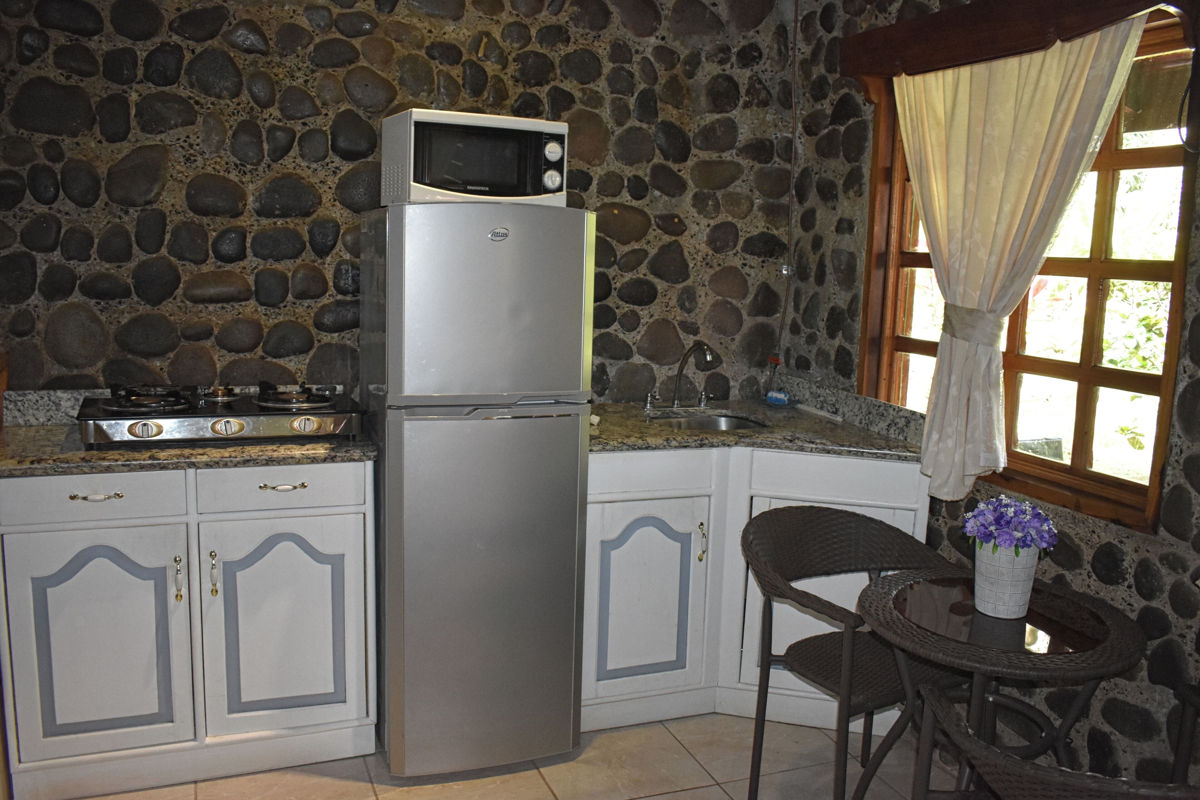 A kitchen with a stone wall, a refrigerator, a microwave, a stove, and a dining area with a chair and table.