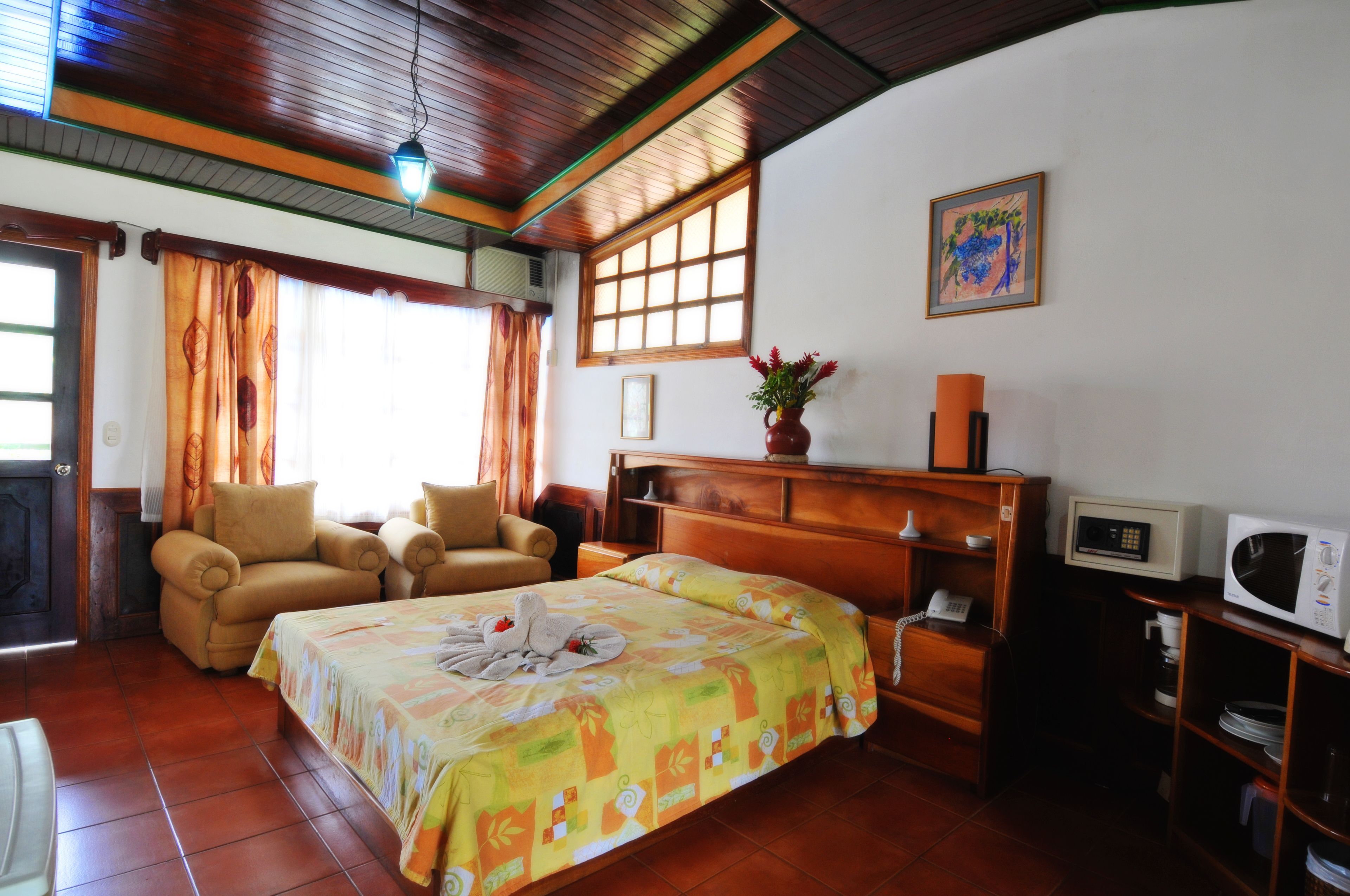 A hotel room with a bed, two chairs, a wooden dresser, a microwave, and a potted plant.