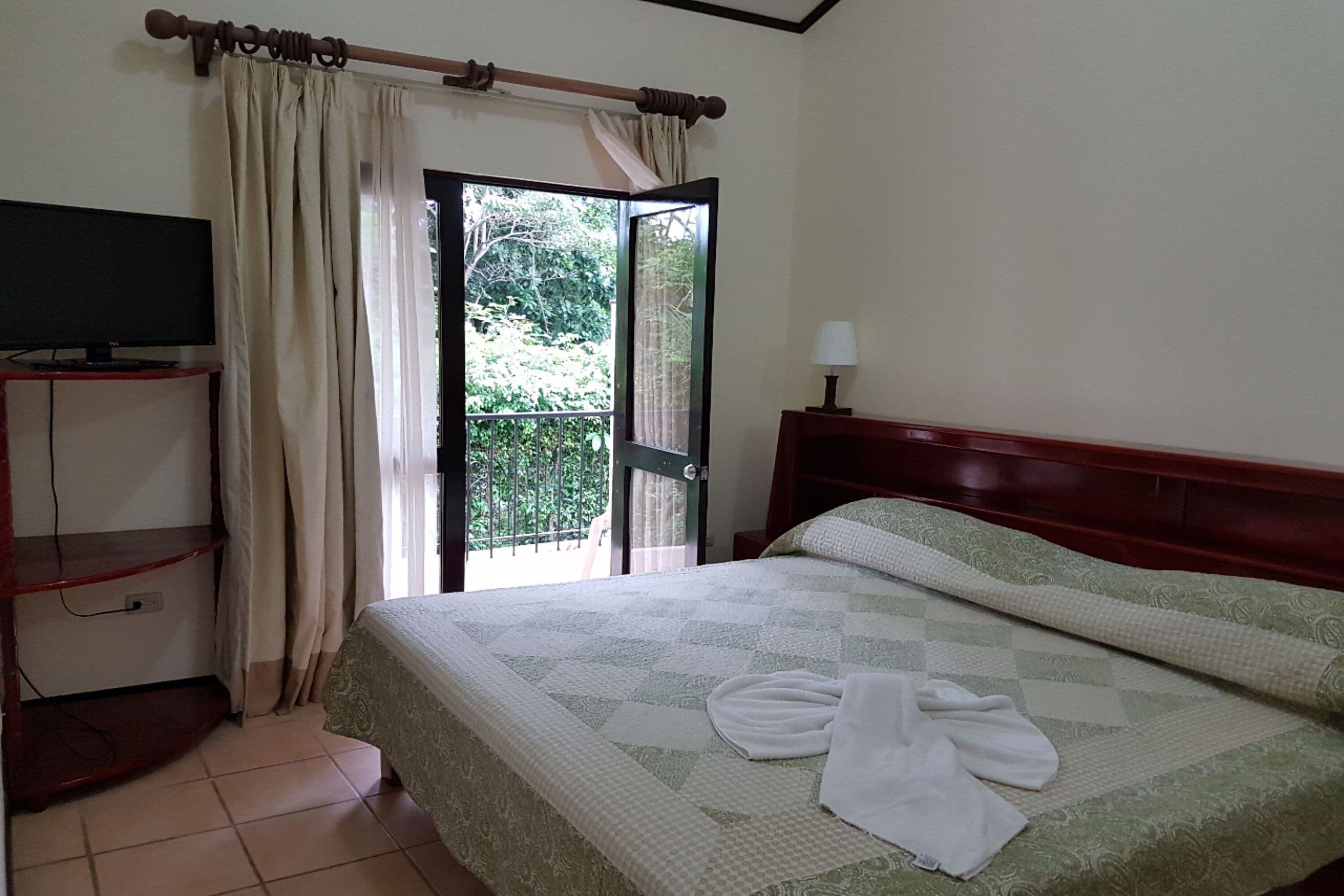 A hotel room with a bed, a TV on a stand, a balcony with curtains, and a lamp.