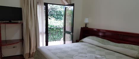 A hotel room with a bed, a TV on a stand, a balcony with curtains, and a lamp.