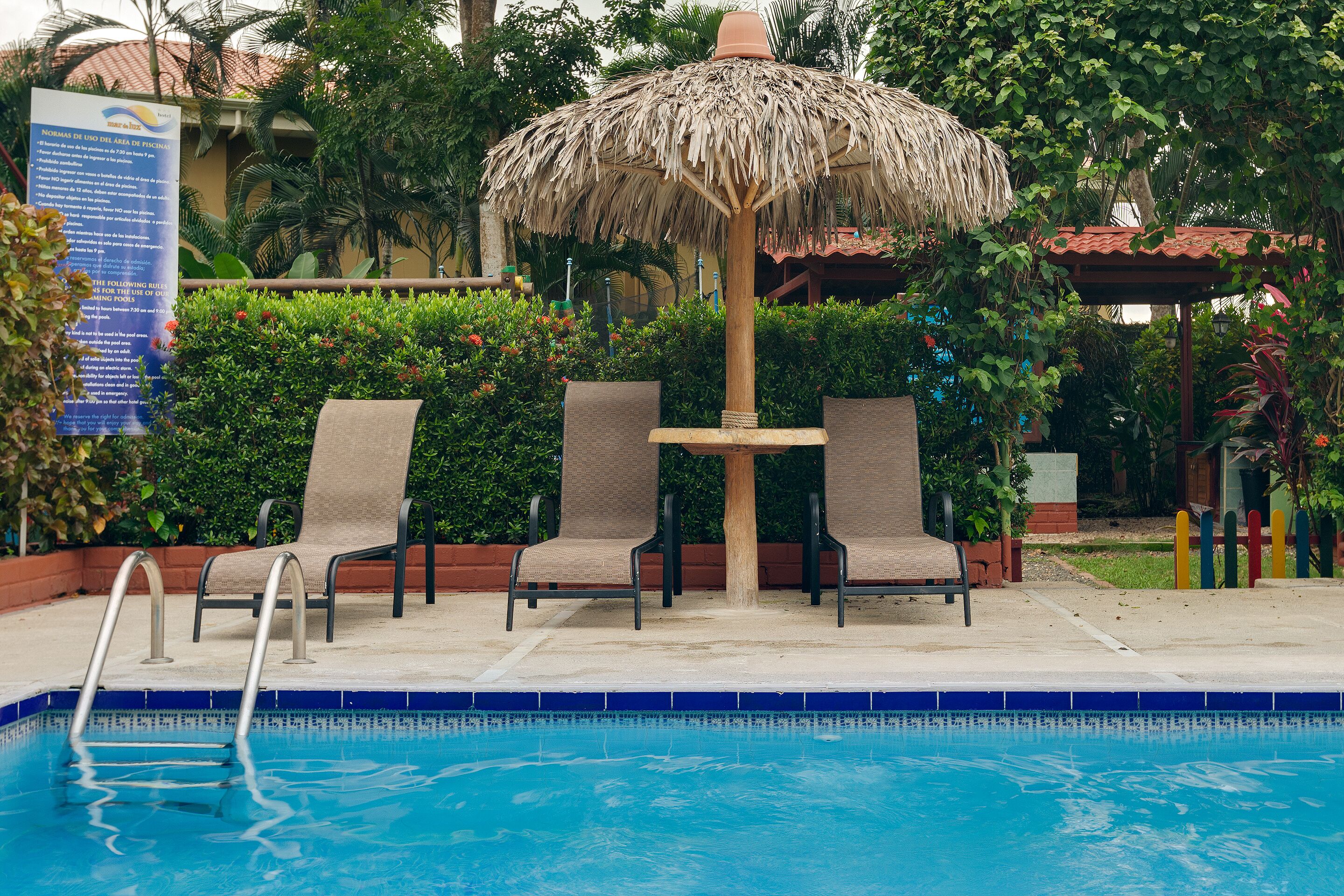 Un'area piscina con un ombrellone di paglia, due poltrone da relax e un cartello.