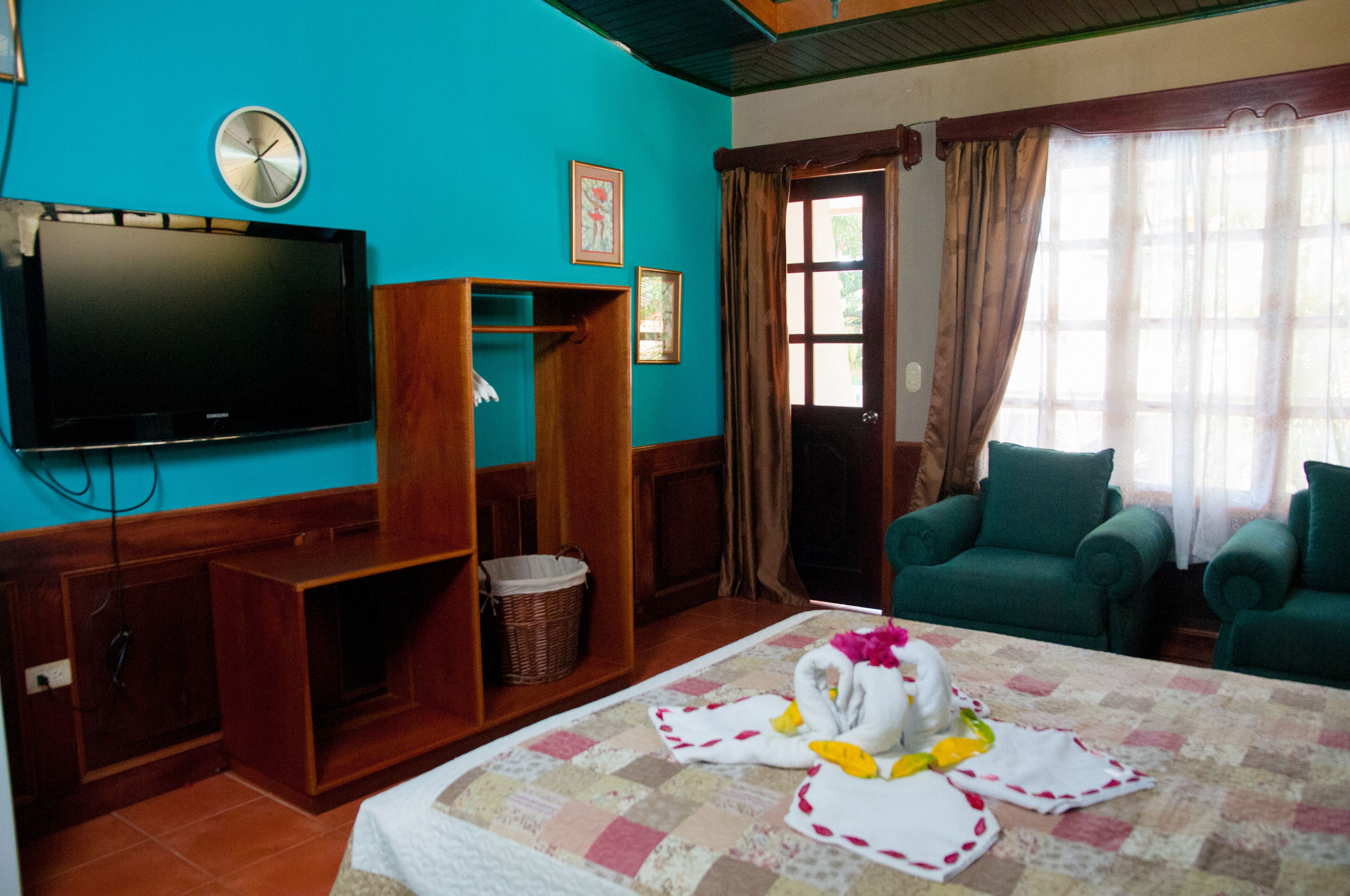 A room with a television, a wooden shelf, a green armchair, a bed with a quilt, and a window with curtains.