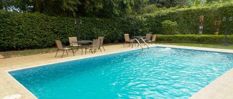 A rectangular swimming pool with a tiled edge, surrounded by a hedge and chairs.