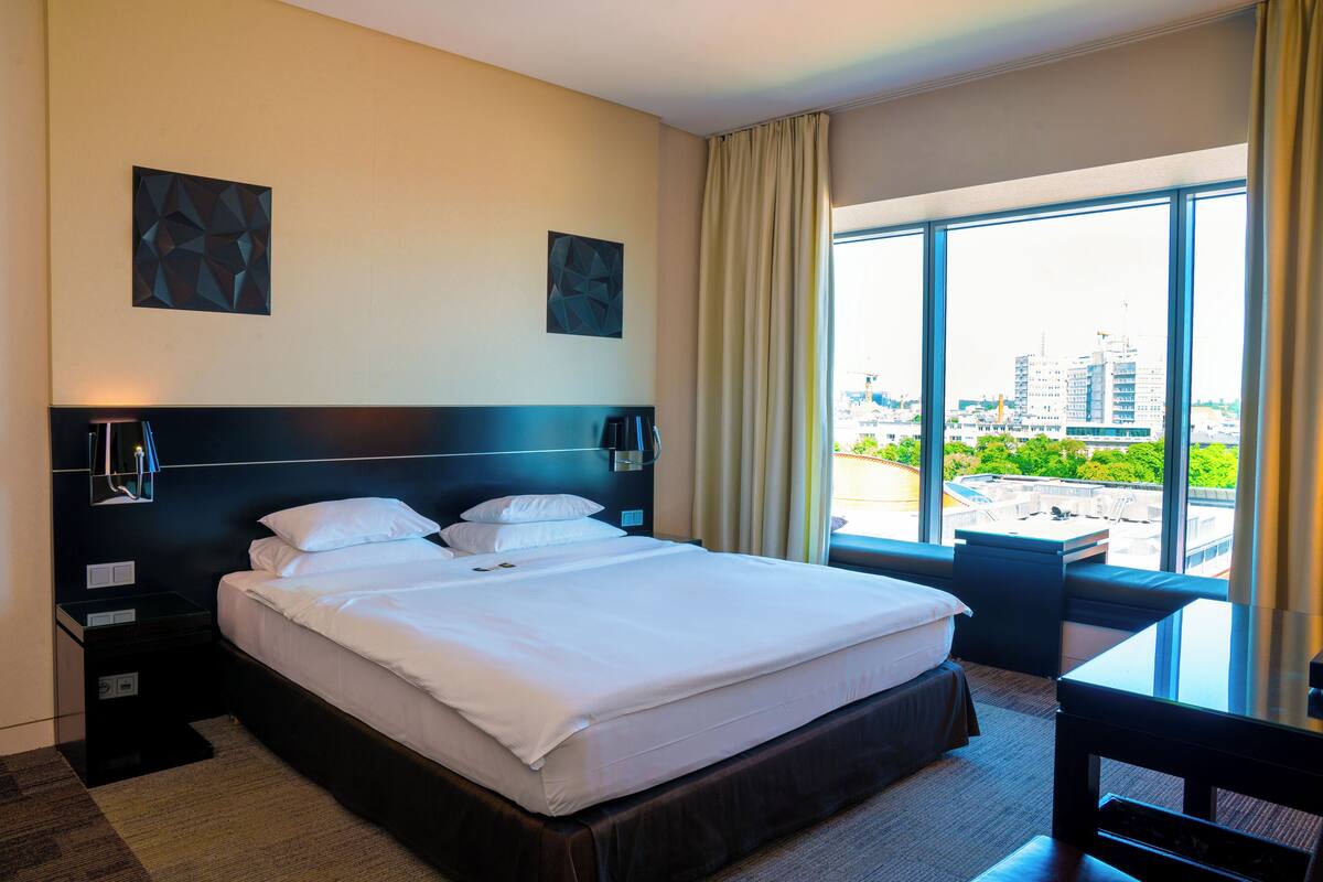 Habitación de hotel con una cama grande, un escritorio y vistas de la ciudad a través de amplias ventanas.