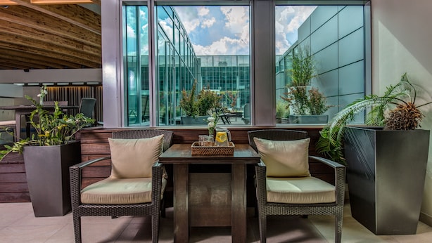 Un espacio al aire libre moderno con sillas de mimbre y una mesa, grandes ventanales con vistas a un edificio y al cielo, y plantas en macetas.