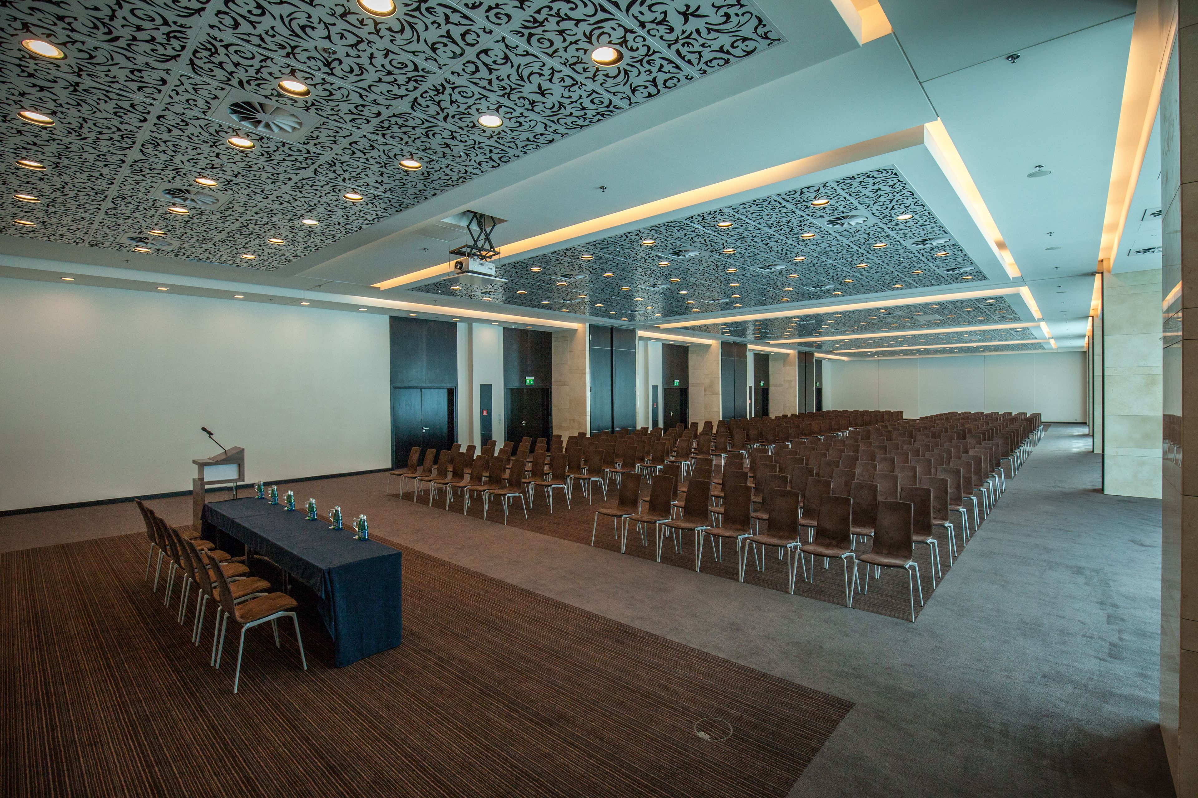 Konferenssihuone, jossa on riveissä tuoleja, pitkä pöytä tuoleineen ja kuvioitu katto.
