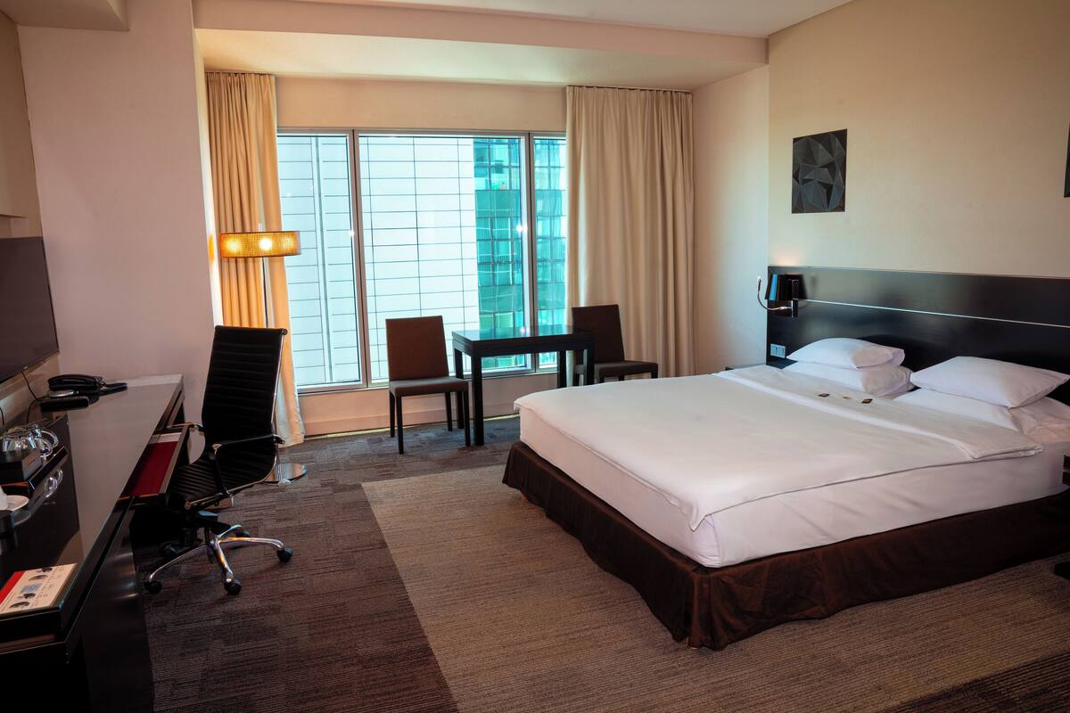 Habitación de hotel con una cama grande, un escritorio con silla, un televisor y una ventana con cortinas.