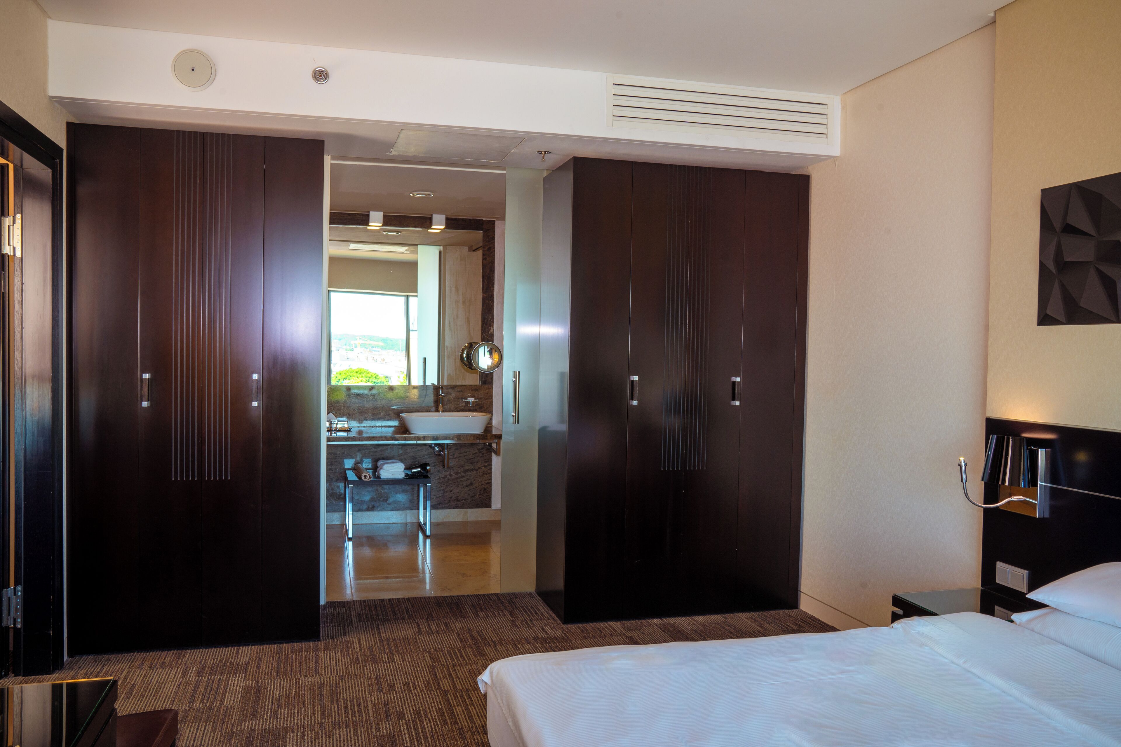 Een hotelkamer met een groot bed, een donkere houten garderobe, een badkamer met een wastafel en spiegel, en uitzicht op buiten.