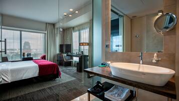 A hotel room with a large bed, a desk, a computer, a bathroom with a large sink and mirror, and a view of a cityscape.