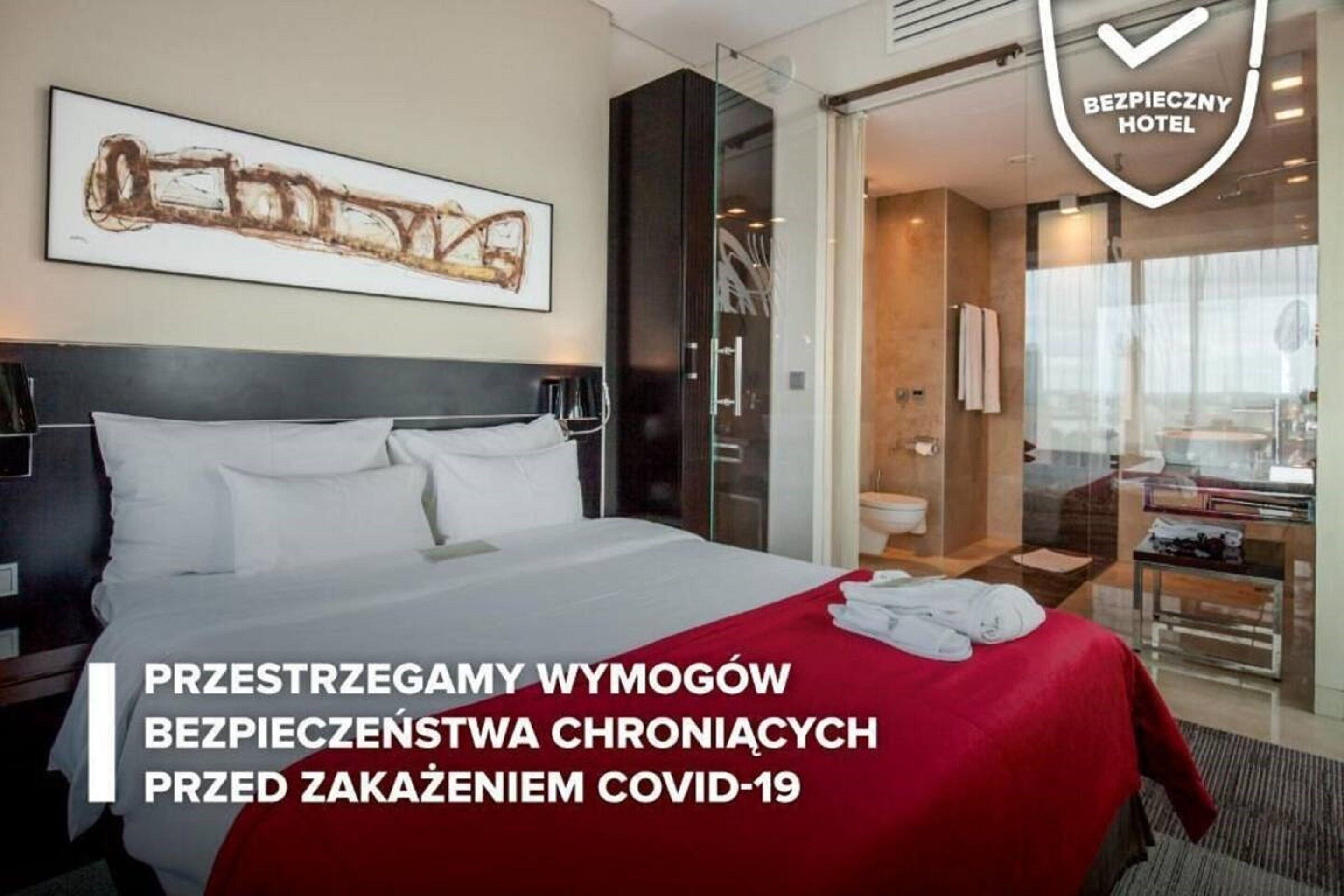 A hotel room with a large bed, a glass door leading to a bathroom, and a framed artwork on the wall.