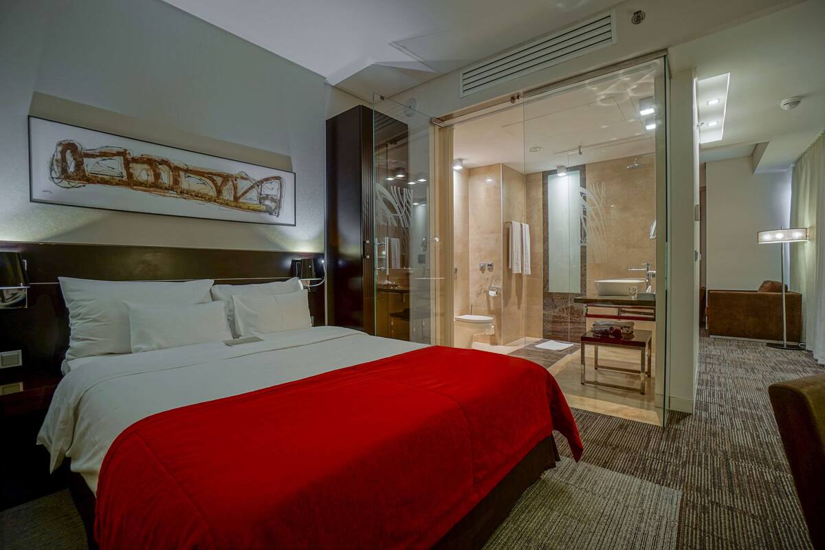 Habitación de hotel con una cama grande, cabina de ducha de vidrio, baño con lavamanos y espejo, y un cuadro en la pared.