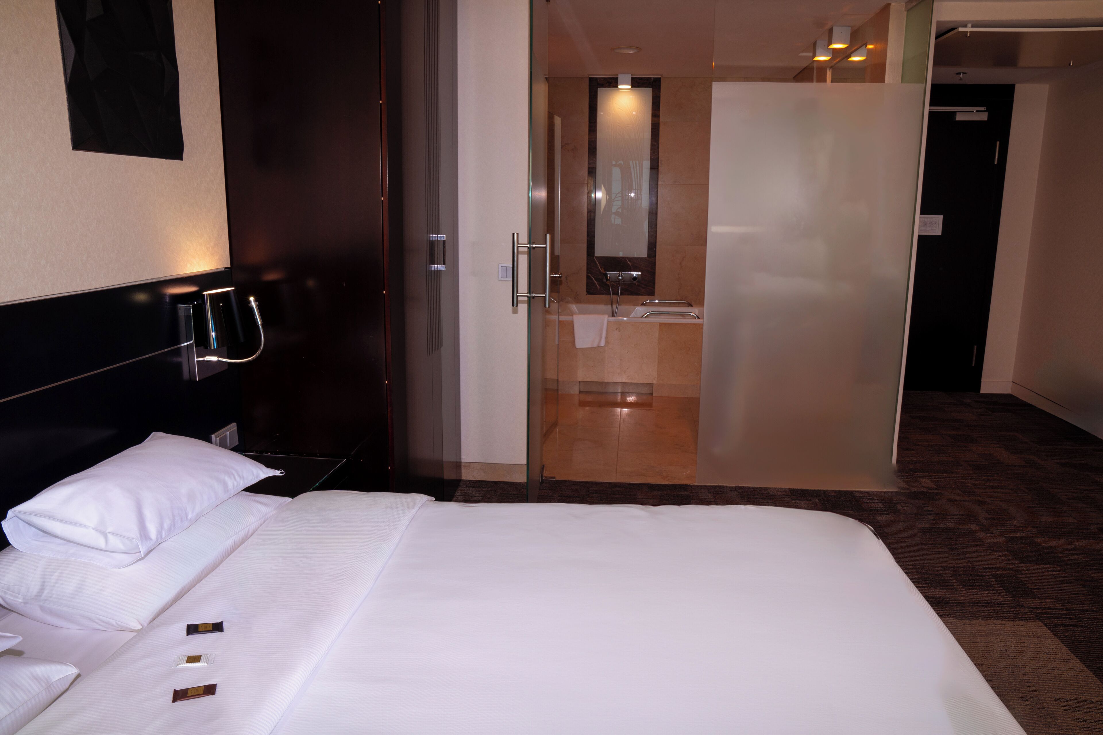 A hotel room with a bed, a bathroom with a glass door, and a mirror.