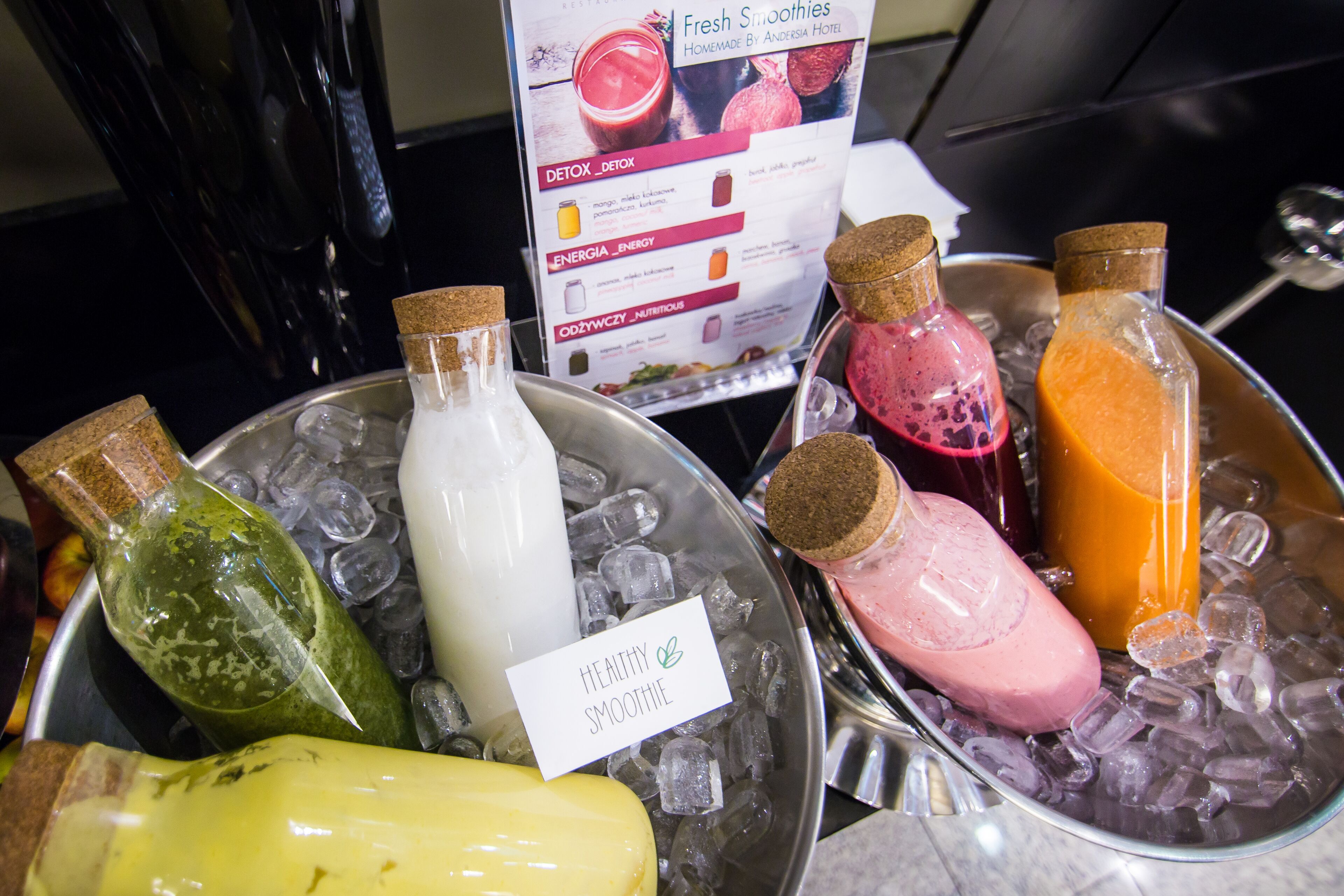 Variedad de batidos en un recipiente metálico con hielo, un menú al fondo y una botella de jugo de naranja con tapón de corcho.