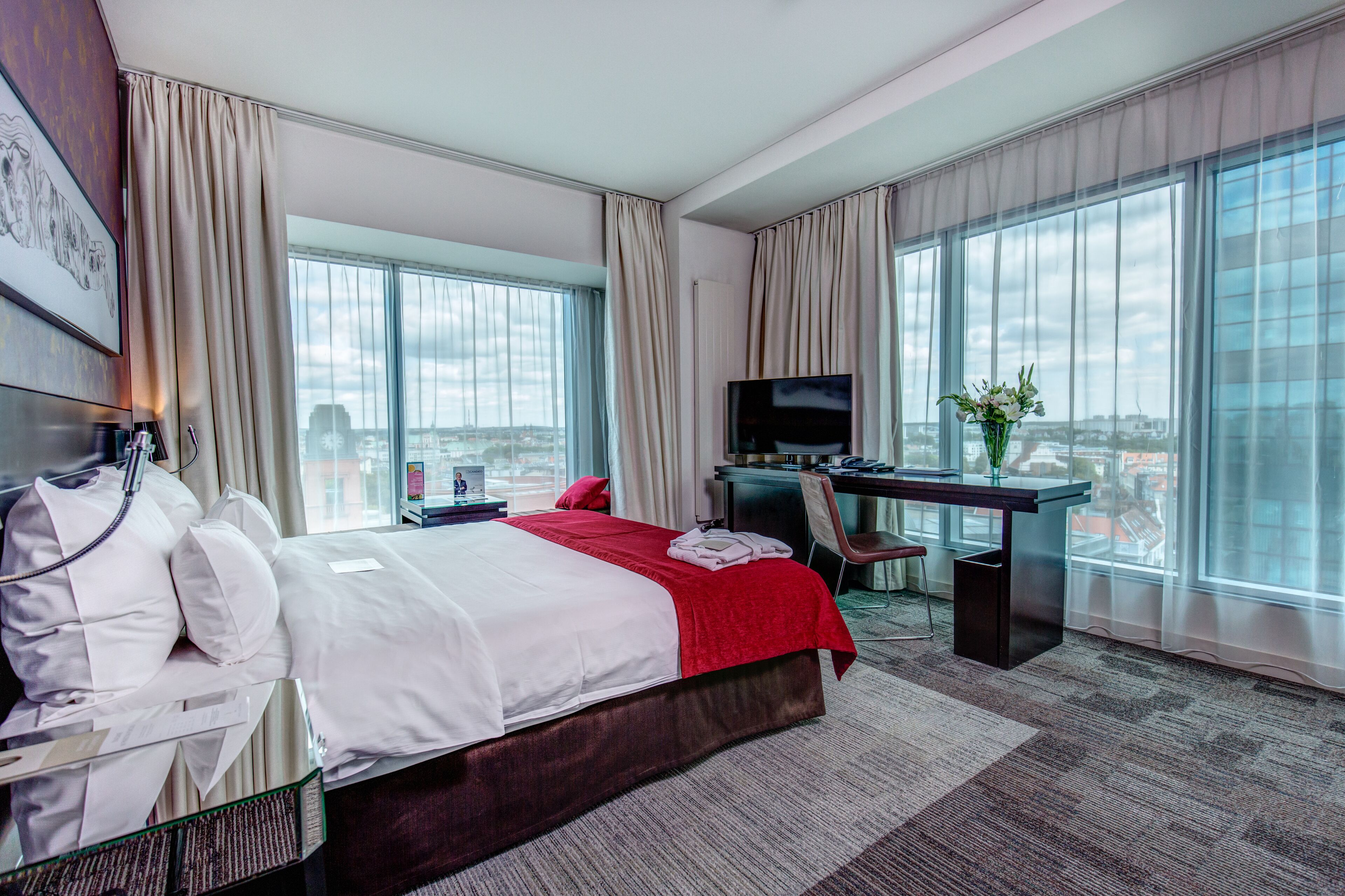 A hotel room with a large bed, a desk with a TV, and a city view.
