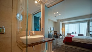 Un baño de hotel moderno con un lavamanos grande, una cabina de ducha de vidrio y un televisor visible en la habitación contigua.