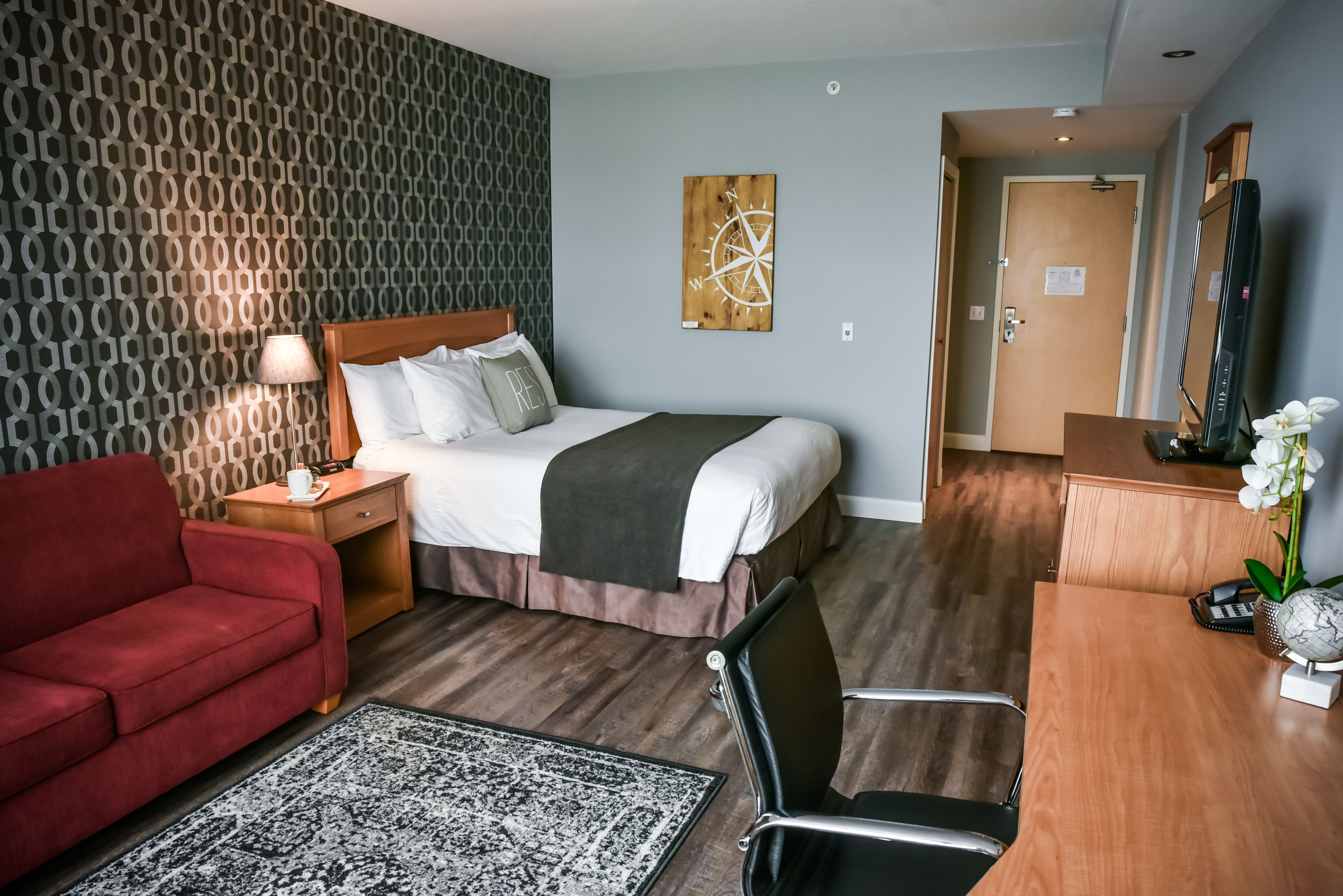 Habitación de hotel con una cama, un sofá rojo, un escritorio con espejo y un cuadro de una brújula en la pared.