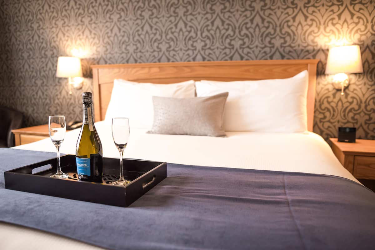 Una cama bien tendida con cabecera de madera, dos lámparas de noche y una bandeja con una botella y vasos sobre la cama.