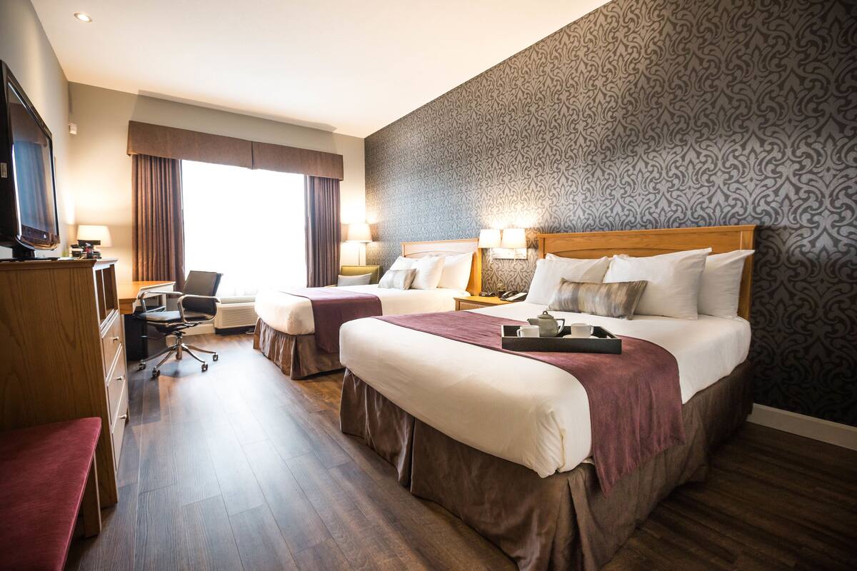 A hotel room with two beds, a desk, a chair, a TV, and a patterned wall.