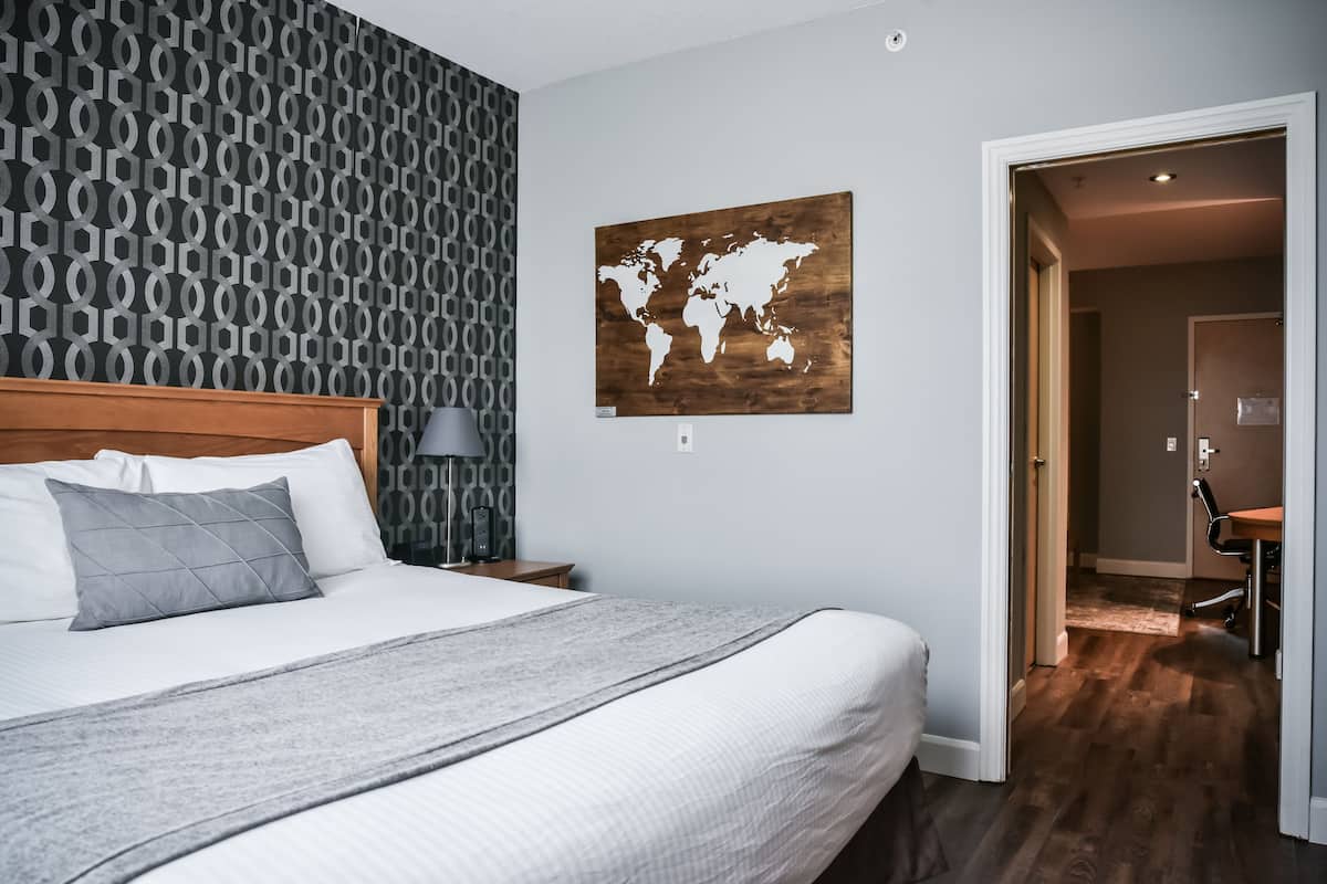 Una cama bien hecha con una manta gris y almohadas, un cuadro de un mapa mundial en la pared y piso de madera.