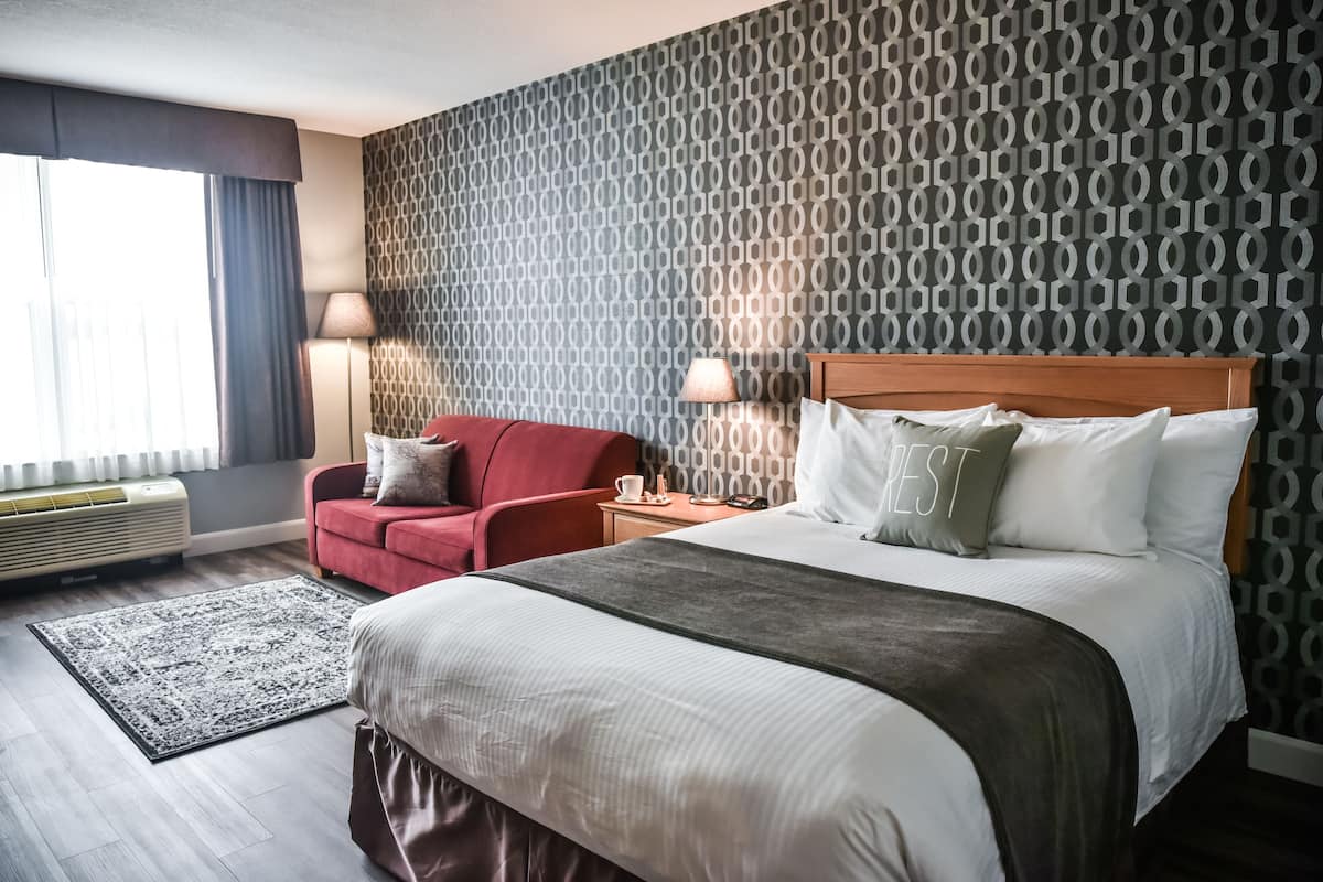 Una habitación de hotel con una cama grande, un sillón rojo, una mesita de noche con lámpara y una pared decorada.