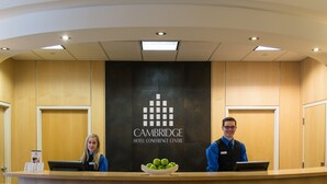A reception desk at the Cambridge Hotel Conference Centre with a logo, two staff members, and a bowl of green apples.
