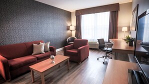 A hotel room with a red sofa, a wooden coffee table, a red armchair, a desk with a chair, a TV, and a large window with curtains.