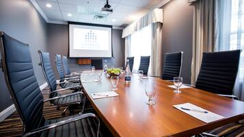 A conference room with a long table, black chairs, a projector screen, and a window with curtains.