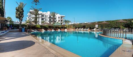 Ein klarer, blauer Pool mit Liegestühlen und Sonnenschirmen, umgeben von einem Gebäude mit Balkonen.