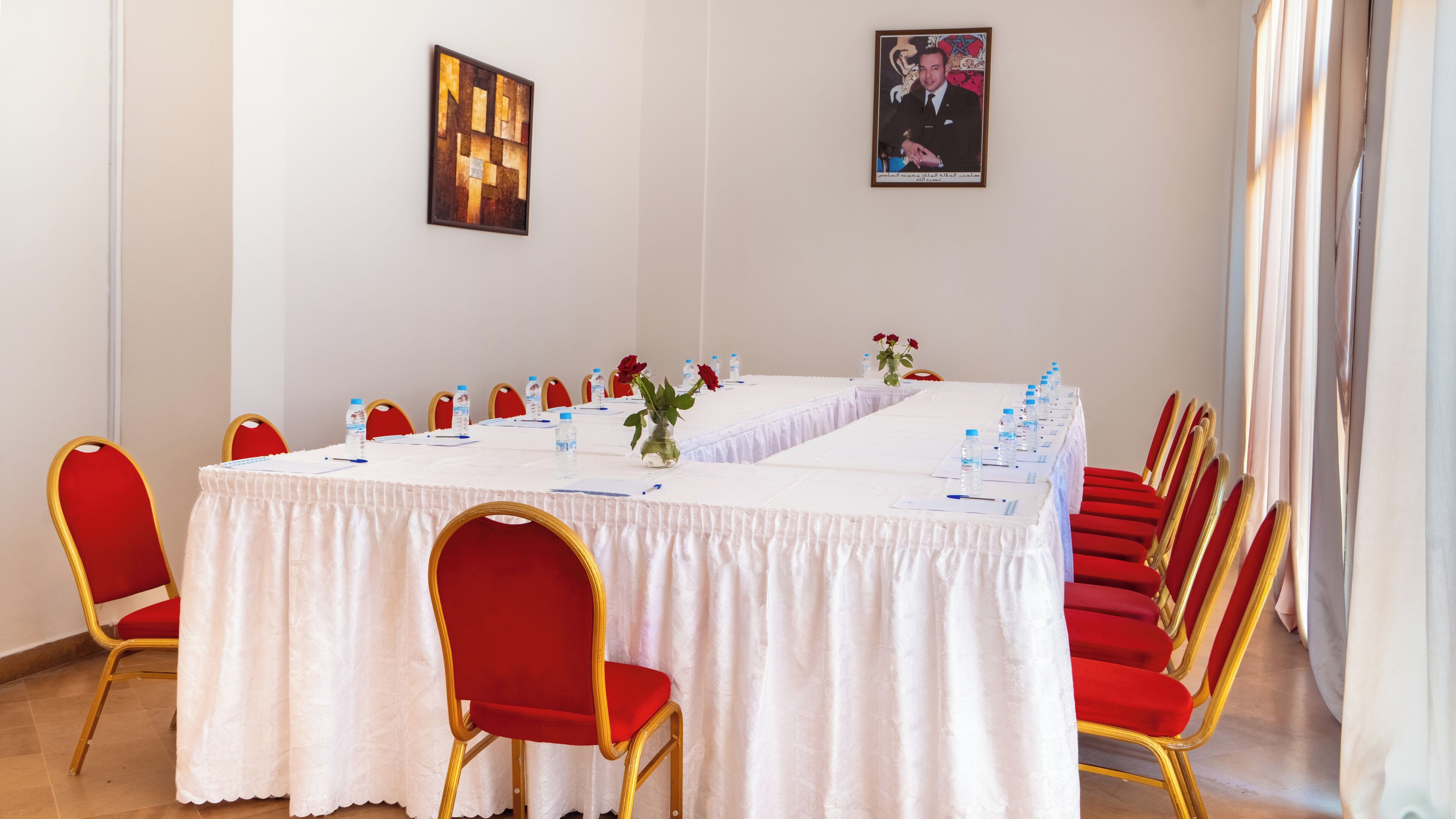 Un salón de conferencias con una larga mesa, sillas rojas, botellas de agua y un florero con flores.
