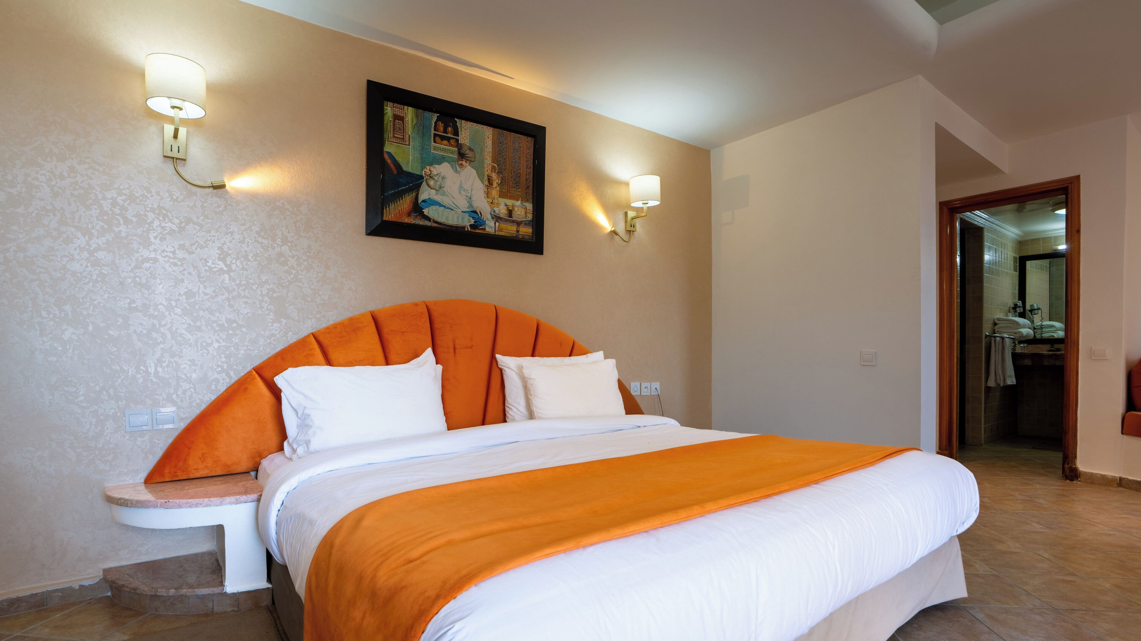 Habitación de hotel con una cama grande, dos lámparas de pared, un cuadro y un baño visible a través de una puerta abierta.