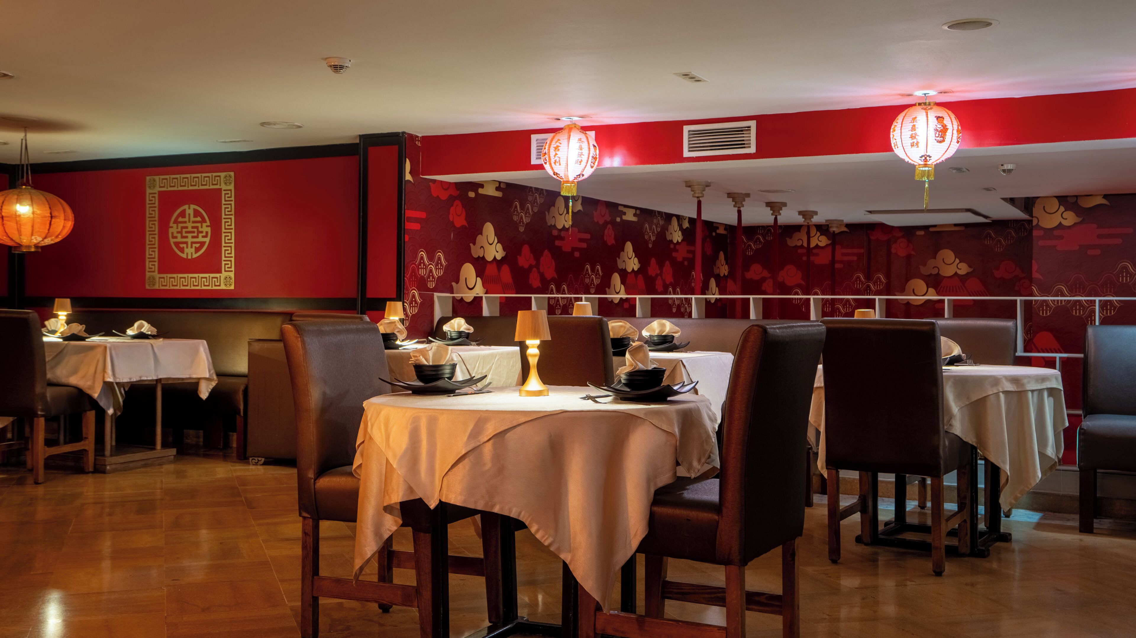 Interior de un restaurante con mesas preparadas para comer, paredes con diseño rojo y faroles colgantes.