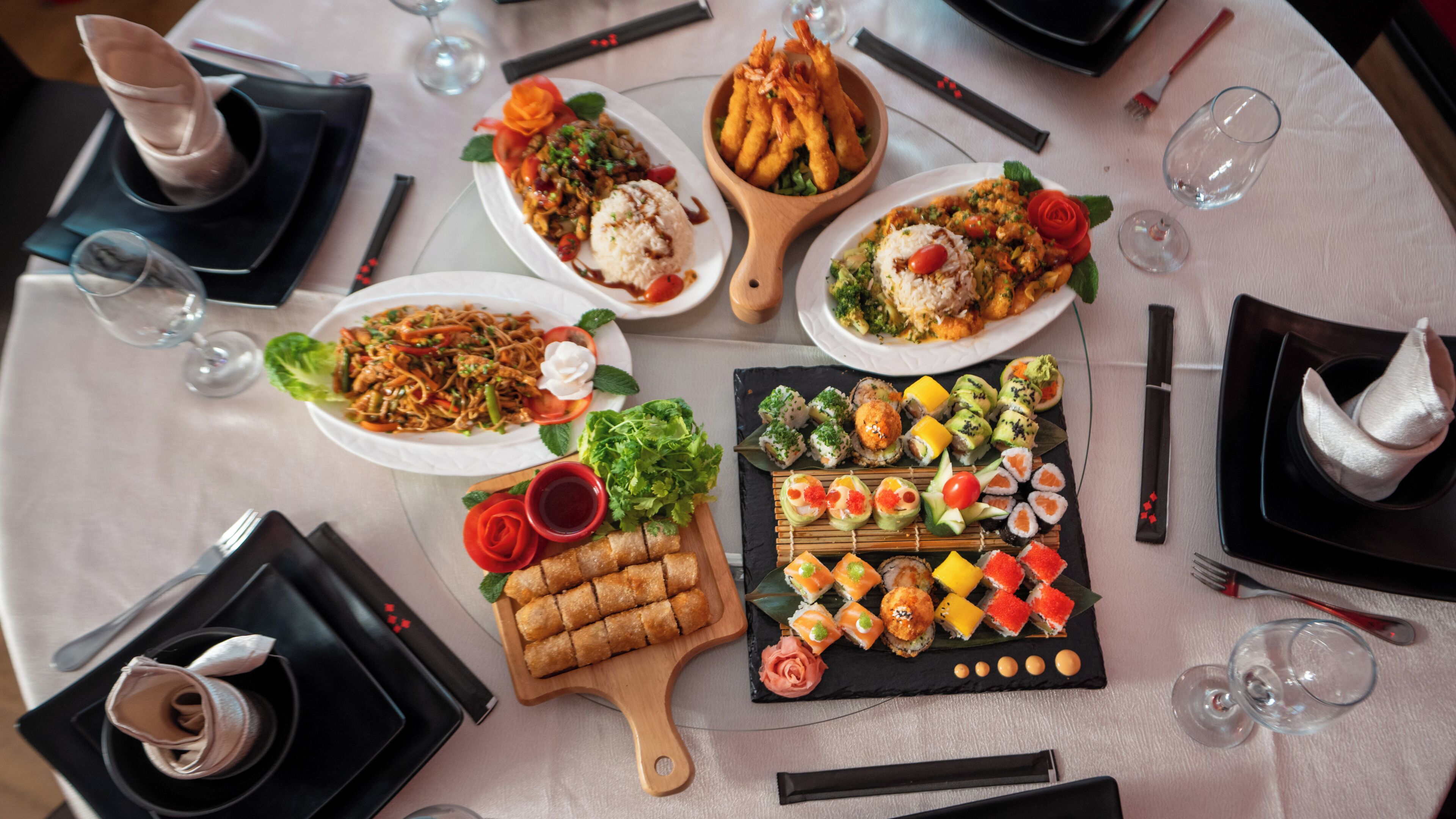 Une table dressée avec divers plats, notamment des sushis, des nouilles et des amuse-gueules, ainsi que des verres à vin et des couverts.