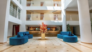 Eine Hotelhalle mit einer Rezeption, zwei blauen Sofas, einer Zierpflanze und einer hohen Decke mit einem Balkon.