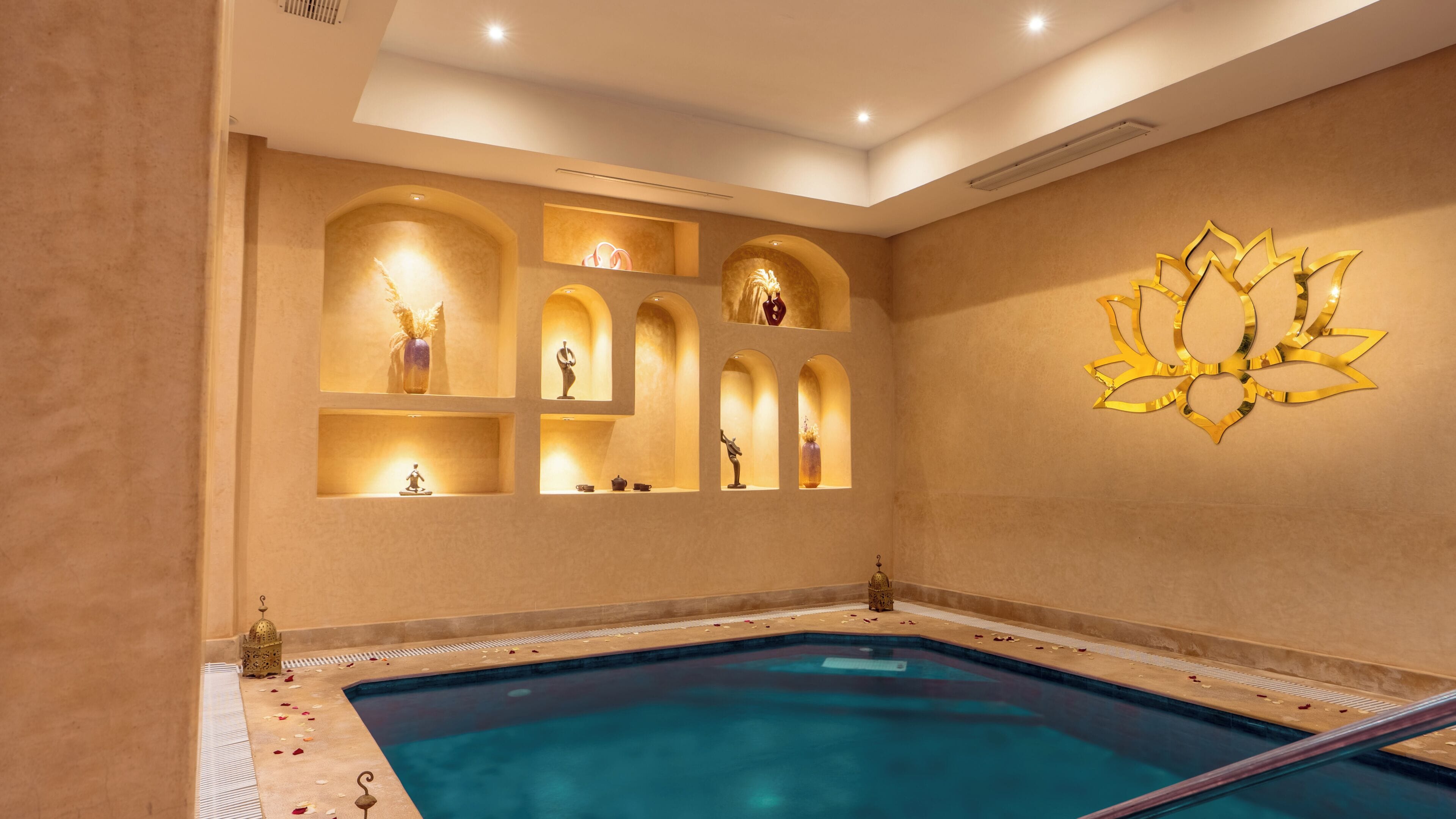 Une piscine à l’eau d’un bleu cristallin, entourée d’un mur beige orné de plusieurs niches en arc abritant des objets décoratifs.