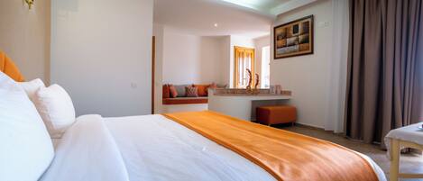 Quarto de hotel com uma cama grande, área de estar com sofá e almofadas, uma pequena mesa e um quadro na parede.