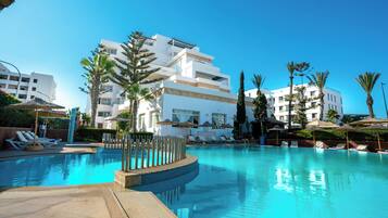 A swimming pool with lounge chairs and umbrellas, surrounded by palm trees and modern buildings.
