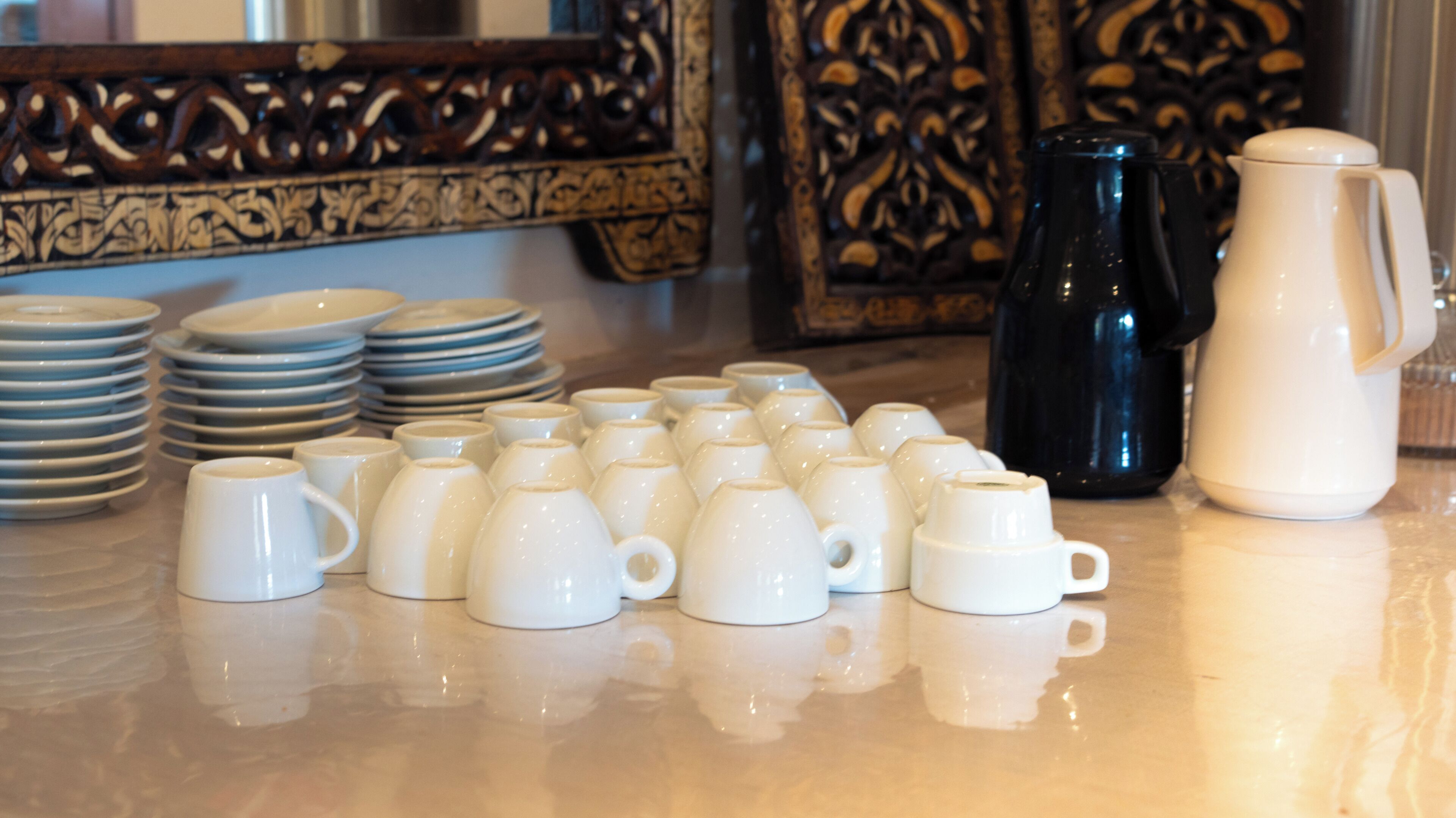 Una colección de tazas y platos de cerámica blanca dispuestos sobre una superficie reflectante, con una cafetera blanco y negro y un panel de madera decorativo al fondo.