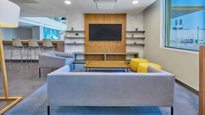 A modern lounge area with a grey sofa, yellow ottoman, and a wooden coffee table. There are bar stools and a flat-screen TV mounted on a wooden wall. - City Express by Marriott Mexicali (Mexicali)
