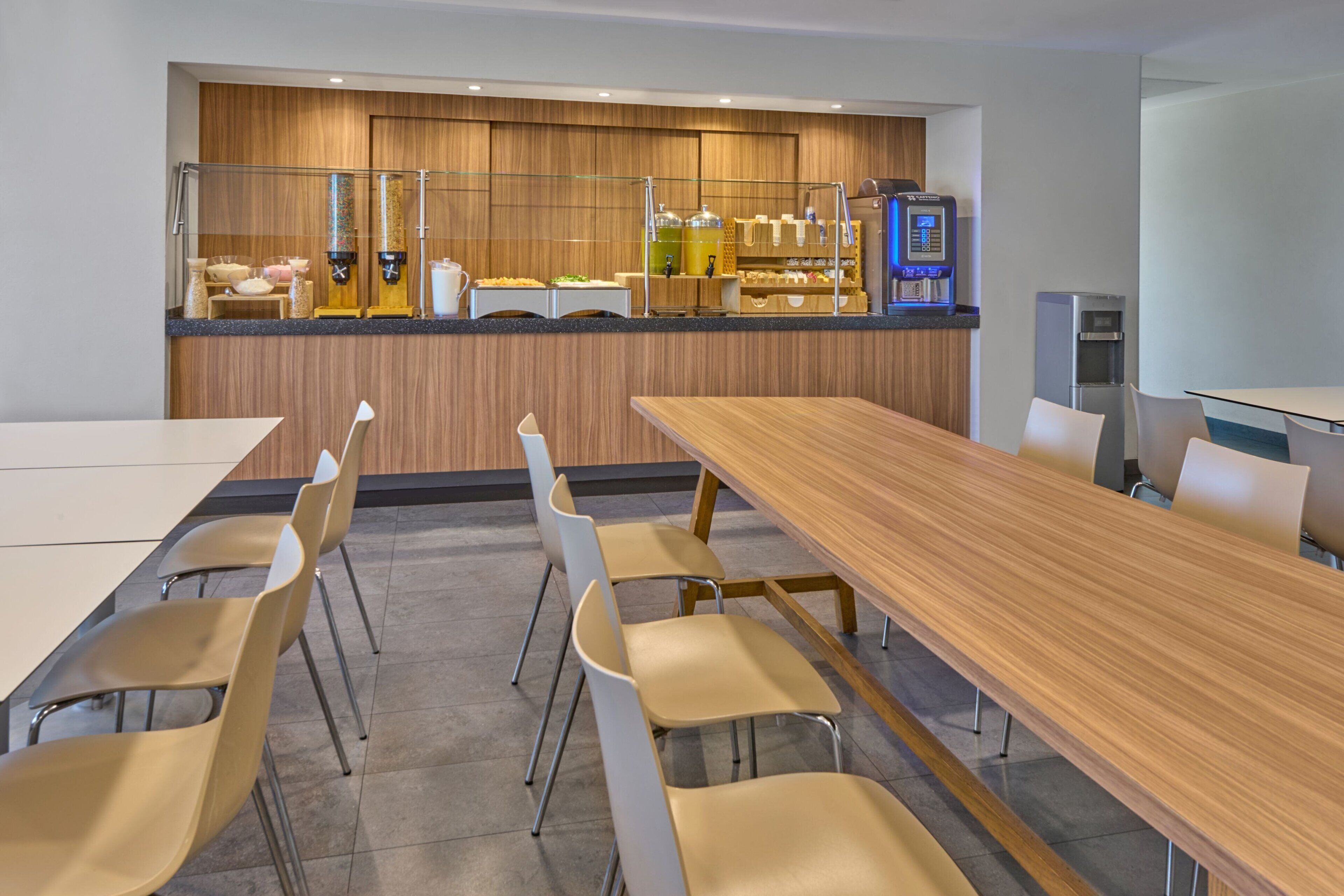 A modern conference room with a long wooden table and beige chairs, a buffet area with food dispensers, and a refrigerator.