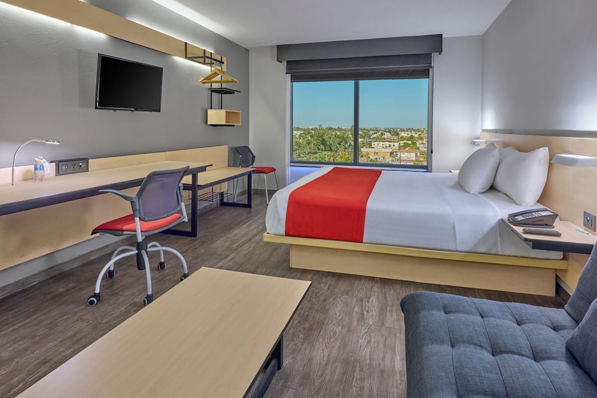 Una habitación de hotel moderna con cama, escritorio, silla y un ventanal con vistas a la ciudad.